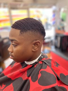 Profile of a client in a red and black cape: waves on top, a low taper at the temple and a sharp line-up at the front.